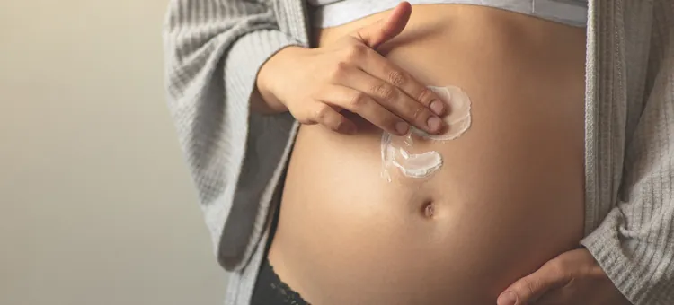 Kobieta w ciąży nakłada krem na swój brzuch w ramach zapobiegania rozstępom, lewą ręką dotyka brzucha