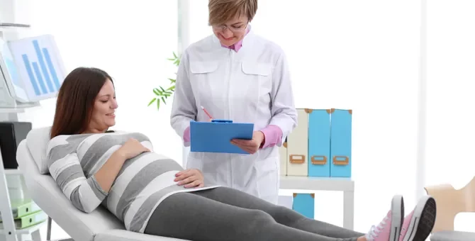 Uśmiechnięta kobieta w ciąży na łóżku w gabinecie oraz lekarka analizująca wyniki
