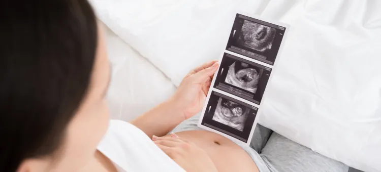 Kobieta w ciąży siedzi na łóżku, trzymając zdjęcie ultrasonograficzne dziecka, prawa ręka dotyka brzucha