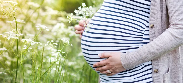 Kobieta w ciąży w pasiastej bluzce stoi na łące wśród kwiatów, trzyma rękę na brzuchu oraz jasne kwiatki, zbliżenie na brzuch