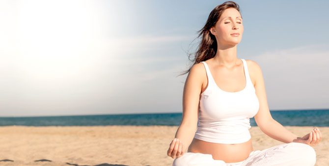 Kobieta w ciąży medytuje na plaży, siedząc w pozycji jogi