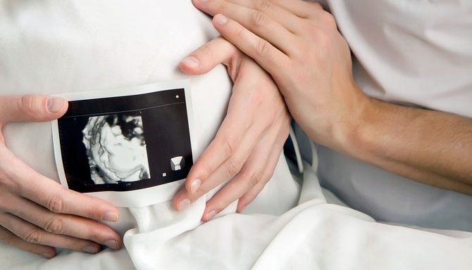 Zbliżenie rąk kobiety trzymającej zdjęcie USG na brzuchu ciążowym, ręka mężczyzny dotyka jej dłoni