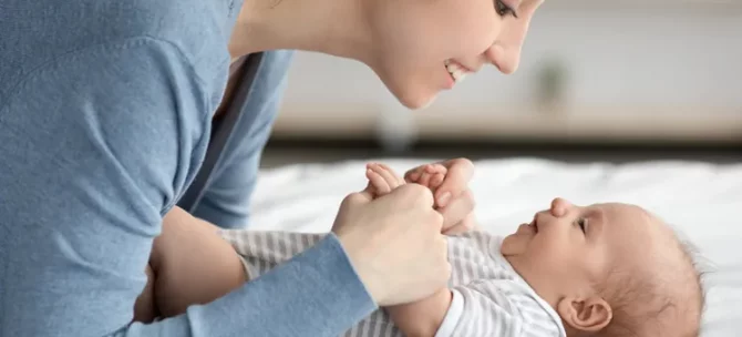 Uśmiechnięta mama w niebieskiej bluzce trzyma za ręce swojego leżącego maluszka