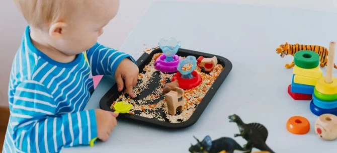 Dziecko bawiące się ziarnami i figurkami na tacy sensorycznej