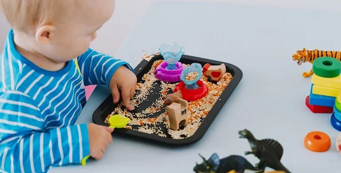 Dziecko bawiące się ziarnami i figurkami na tacy sensorycznej