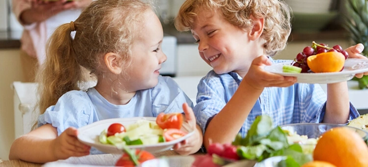 Dwoje uśmiechniętych dzieci je warzywa i owoce – zdrowa, zbilansowana dieta wspierająca odporność dziecka
