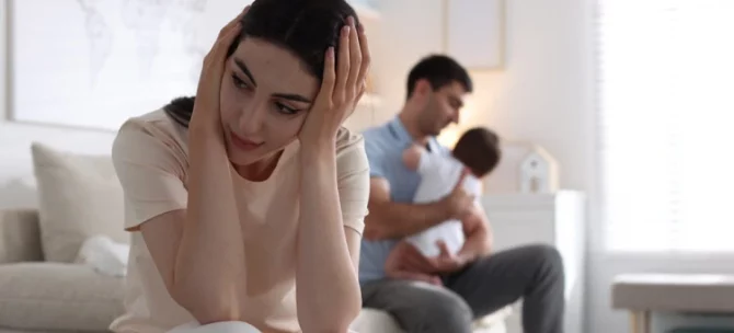 Młoda mama siedząca ze spuszczoną głową, odczuwająca trudne emocje w okresie połogu, w tle tata z niemowlęciem