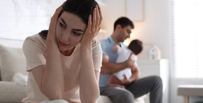 Młoda mama siedząca ze spuszczoną głową, odczuwająca trudne emocje w okresie połogu, w tle tata z niemowlęciem