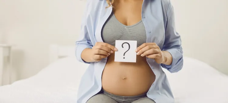 Młoda kobieta w ciąży, siedząca na łóżku, trzymająca kartkę z pytajnikiem, zastanawiająca się nad imieniem dla dziecka