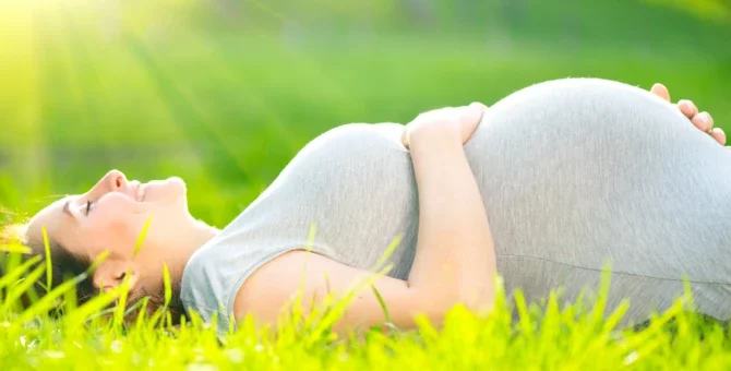 Szczęśliwa kobieta w ciąży dotyka swojego brzucha, leżąc na zielonej trawie i ciesząc się naturą