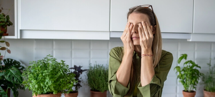 Zmęczona kobieta zasłaniająca oczy dłońmi, siedząca w domowej kuchni