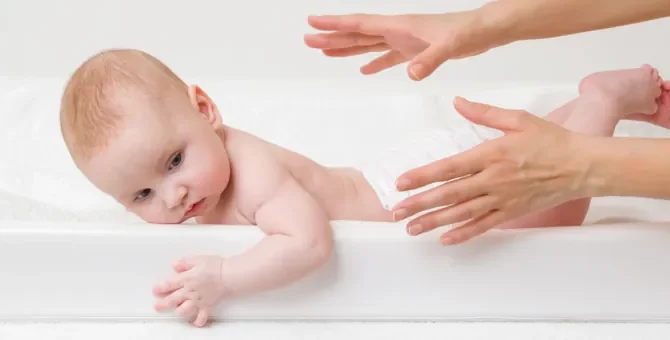 Ręce młodej matki próbują złapać niemowlę, leżące na białym materacu