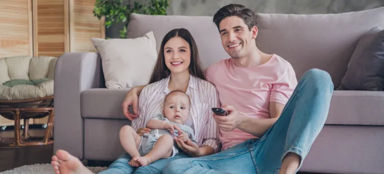 Urocza rodzina ogląda wspólnie telewizję, siedząc na podłodze, opierając się o kanapę, w komfortowym mieszkaniu