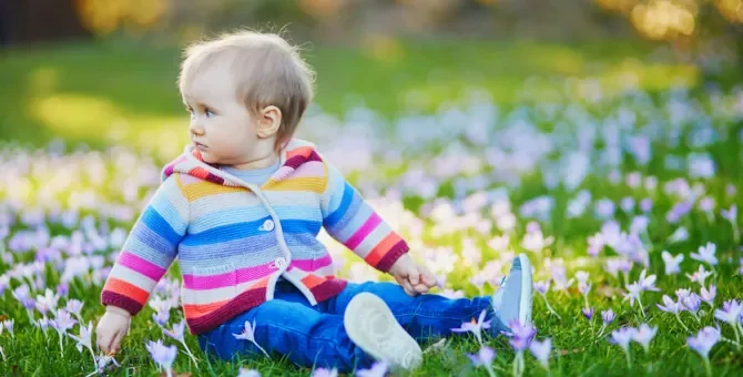 Małe dziecko w kolorowym ubraniu, siedzące na trawie i trzymające fioletowy kwiat podczas słonecznego wiosennego dnia
