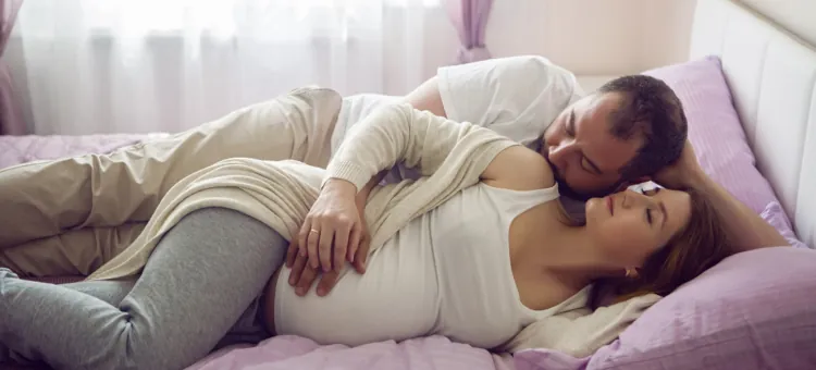 Mąż w białej koszulce całuje w ramię swoją ciężarną żonę, dotyka jej brzucha, oboje leżą na łóżku