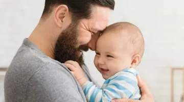 Ojciec w szarej koszulce i syn niemowlak w pasiastym stroju, którzy z radością spędzają razem czas w domu, przytulają się