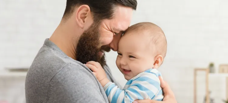 Ojciec w szarej koszulce i syn niemowlak w pasiastym stroju, którzy z radością spędzają razem czas w domu, przytulają się