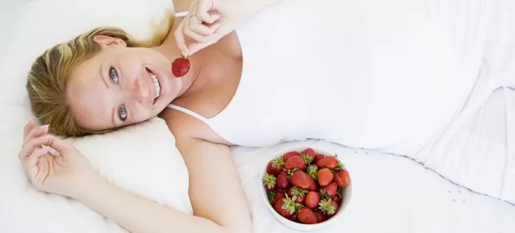 Ciężarna kobieta w białym ubraniu leży na białym łóżku i uśmiecha się, trzymając truskawkę, obok niej miska pełna truskawek