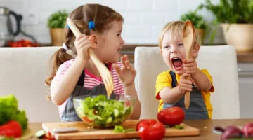 Dzieci w trakcie przygotowania sałatki warzywnej w kuchni, trzymają łyżkę i widelec drewniany, bawią i śmieją się