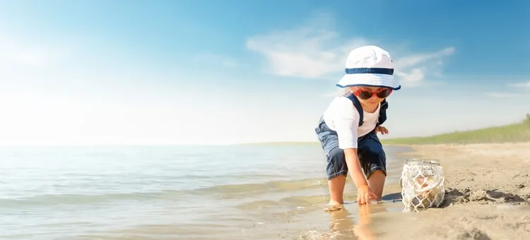 Małe dziecko w kapeluszu bawiące się na brzegu morza, obok stoi przezroczysty słój na muszelki