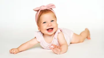 Na białym tle leży rude niemowle dziewczynka, w różowym body i opasce na głowie