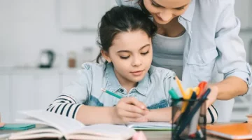 Dziewczynka trzyma ołówek, odrabia lekcje przy biurku z pomocą dorosłej osoby podczas nauczania domowego