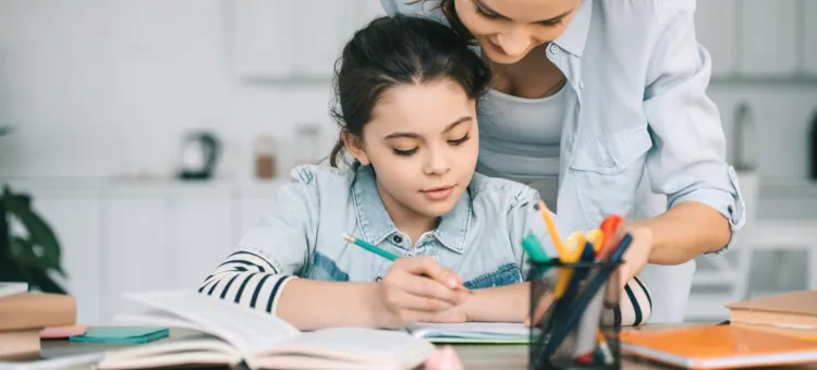 Dziewczynka trzyma ołówek, odrabia lekcje przy biurku z pomocą dorosłej osoby podczas nauczania domowego