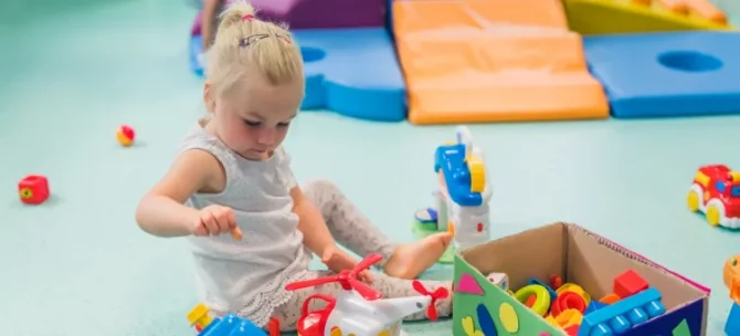 Mała dziewczynka w białej bluzce koncentruje się na zabawie kolorowymi zabawkami na miękkiej, niebieskiej macie w pokoju zabaw