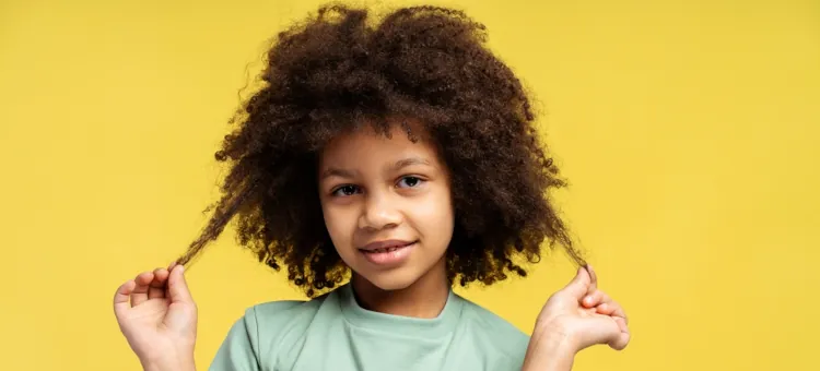 Uśmiechnięta dziewczynka z fryzurą afro bawi się swoimi kręconymi włosami na żółtym tle