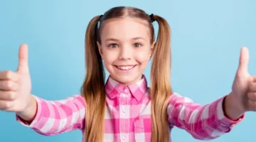 Uśmiechnięta dziewczynka w różowej koszuli w kratę i fryzurą z kucykami pozuje na niebieskim tle, pokazując oba kciuki w geście aprobaty