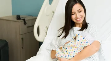 Szczęśliwa kobieta w szpitalnej koszuli czule trzyma swoje nowo narodzone dziecko, leżąc na łóżku