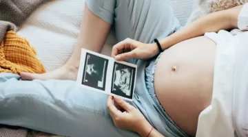 Kobieta w ciąży leży i trzyma w dłoniach i ogląda dwa zdjęcia USG swojego dziecka, zbliżenie na brzuch