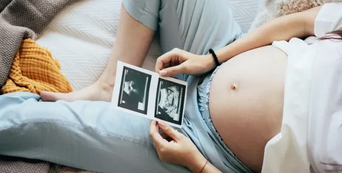 Kobieta w ciąży leży i trzyma w dłoniach i ogląda dwa zdjęcia USG swojego dziecka, zbliżenie na brzuch