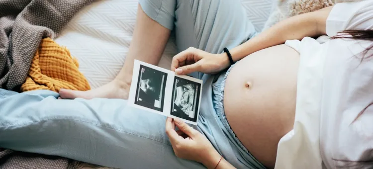 Kobieta w ciąży leży i trzyma w dłoniach i ogląda dwa zdjęcia USG swojego dziecka, zbliżenie na brzuch