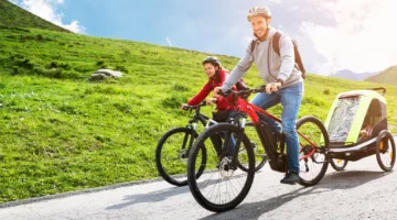 Mężczyzna i kobieta jadą na rowerach górskich po asfaltowej drodze w terenie górzystym z przyczepką przy rowerze mężczyzny