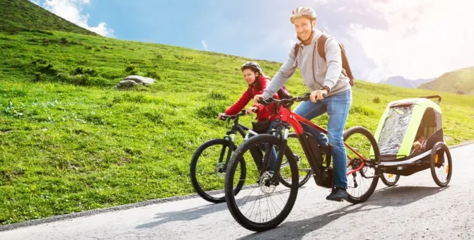 Mężczyzna i kobieta jadą na rowerach górskich po asfaltowej drodze w terenie górzystym z przyczepką przy rowerze mężczyzny