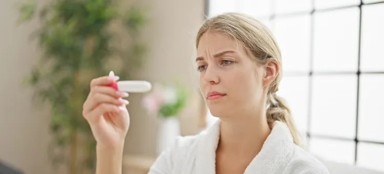 Blondynka w szlafroku trzyma w ręku test ciążowy i patrzy na niego z poważnym wyrazem twarzy