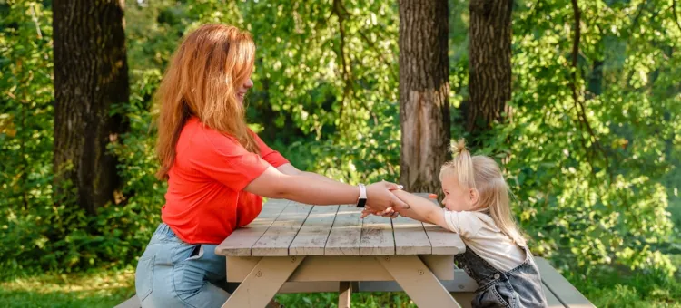 Matka z córką bawią się trzymając się za ręce siedząc na ławkach przy stole w plenerze