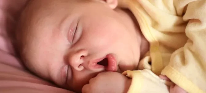 Śpiące niemowlę w żółtym stroju z otwartą buzią leży na różowej pościeli