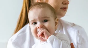 Mama o poważnym wyrazie twarzy trzyma na rękach niemowlę, które wkłada sobie rączkę do buzi w trakcie ząbkowania