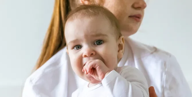Mama o poważnym wyrazie twarzy trzyma na rękach niemowlę, które wkłada sobie rączkę do buzi w trakcie ząbkowania