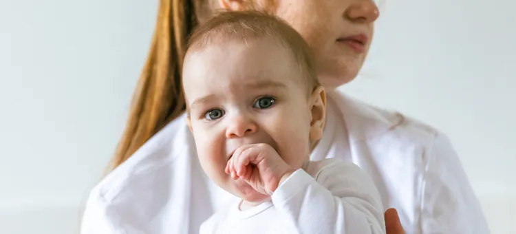 Mama o poważnym wyrazie twarzy trzyma na rękach niemowlę, które wkłada sobie rączkę do buzi w trakcie ząbkowania