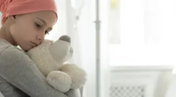 Zamyślone dziecko, chore na białaczkę, w różowej chuście czule obejmuje pluszaka w klinicznych warunkach