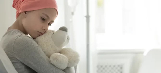 Zamyślone dziecko, chore na białaczkę, w różowej chuście czule obejmuje pluszaka w klinicznych warunkach