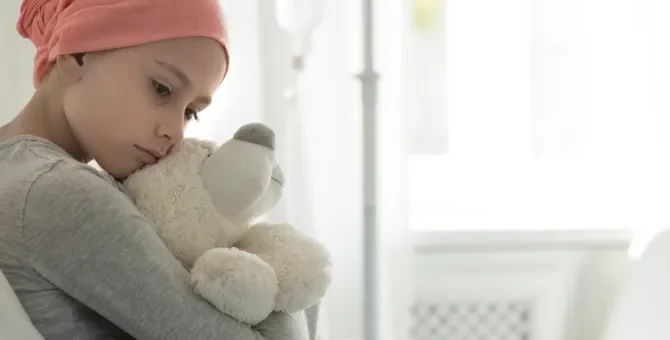 Zamyślone dziecko, chore na białaczkę, w różowej chuście czule obejmuje pluszaka w klinicznych warunkach