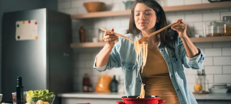 Zadowolona kobieta w ciąży, z uśmiechem wąchająca aromat świeżo ugotowanego spaghetti w kuchni