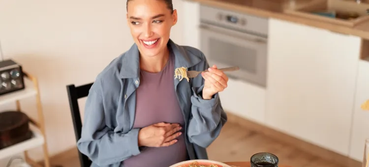 Szczęśliwa przyszła mama z dłonią na brzuchu degustuje posiłek makaronowy w kuchni