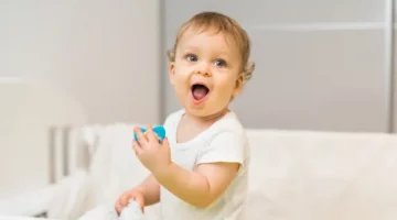 Wesołe dziecko z szeroko otwartymi ustami i oczami trzyma w rączce niebieski smoczek, stojąc w łóżeczku