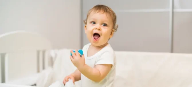 Wesołe dziecko z szeroko otwartymi ustami i oczami trzyma w rączce niebieski smoczek, stojąc w łóżeczku