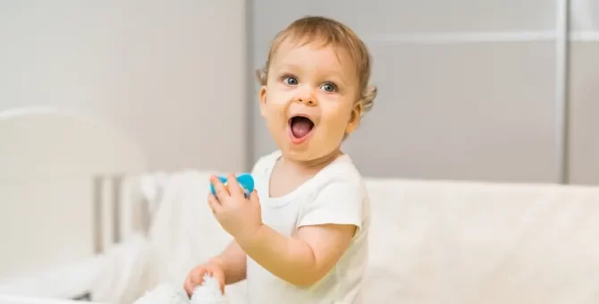 Wesołe dziecko z szeroko otwartymi ustami i oczami trzyma w rączce niebieski smoczek, stojąc w łóżeczku